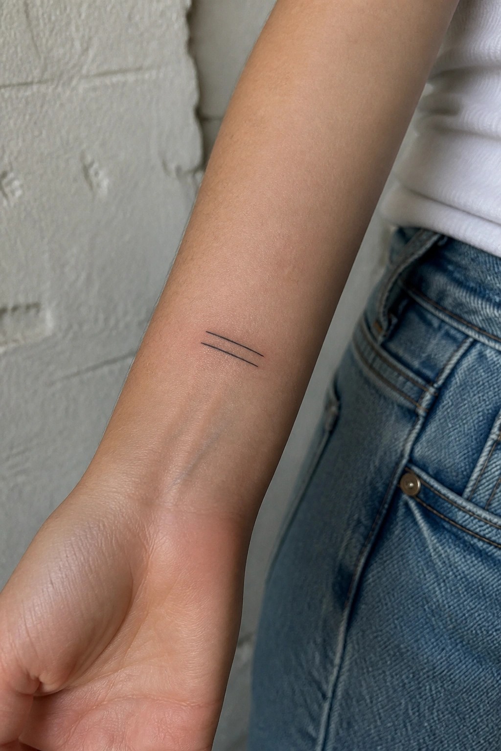 Wrist with two parallel black line tattoo, white shirt, blue jeans, brick wall.
