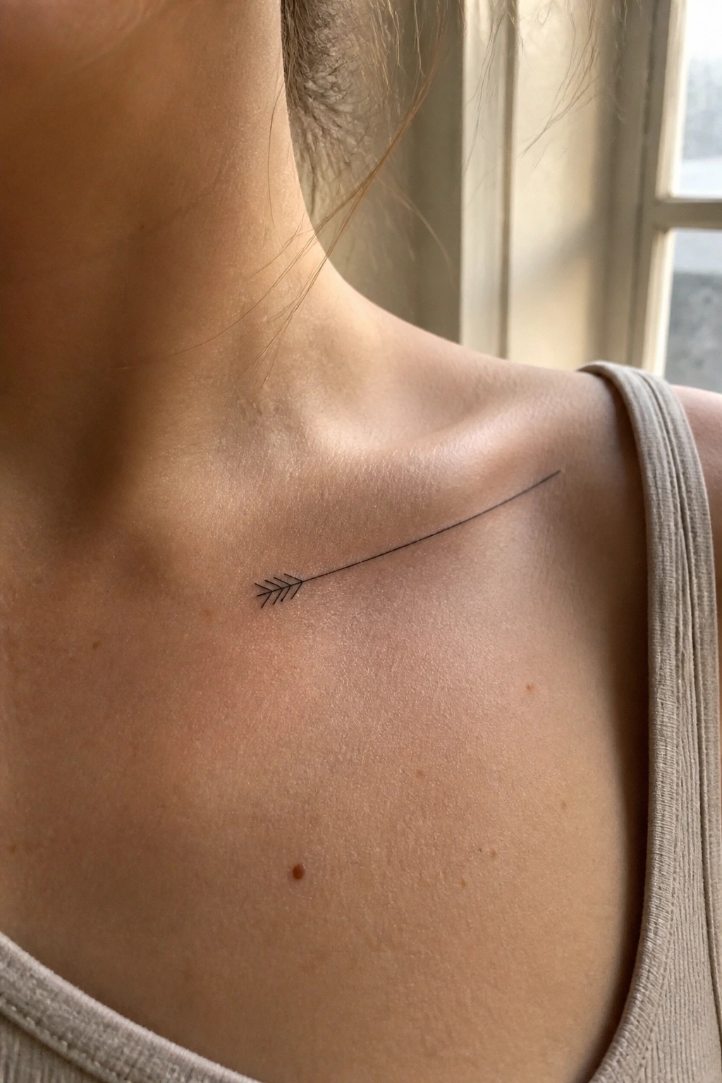 Close-up of a small black arrow tattoo on fair skin near the collarbone, beige strap visible.