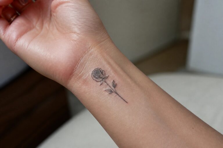Light-skinned wrist with a fine-line black rose tattoo and stem.