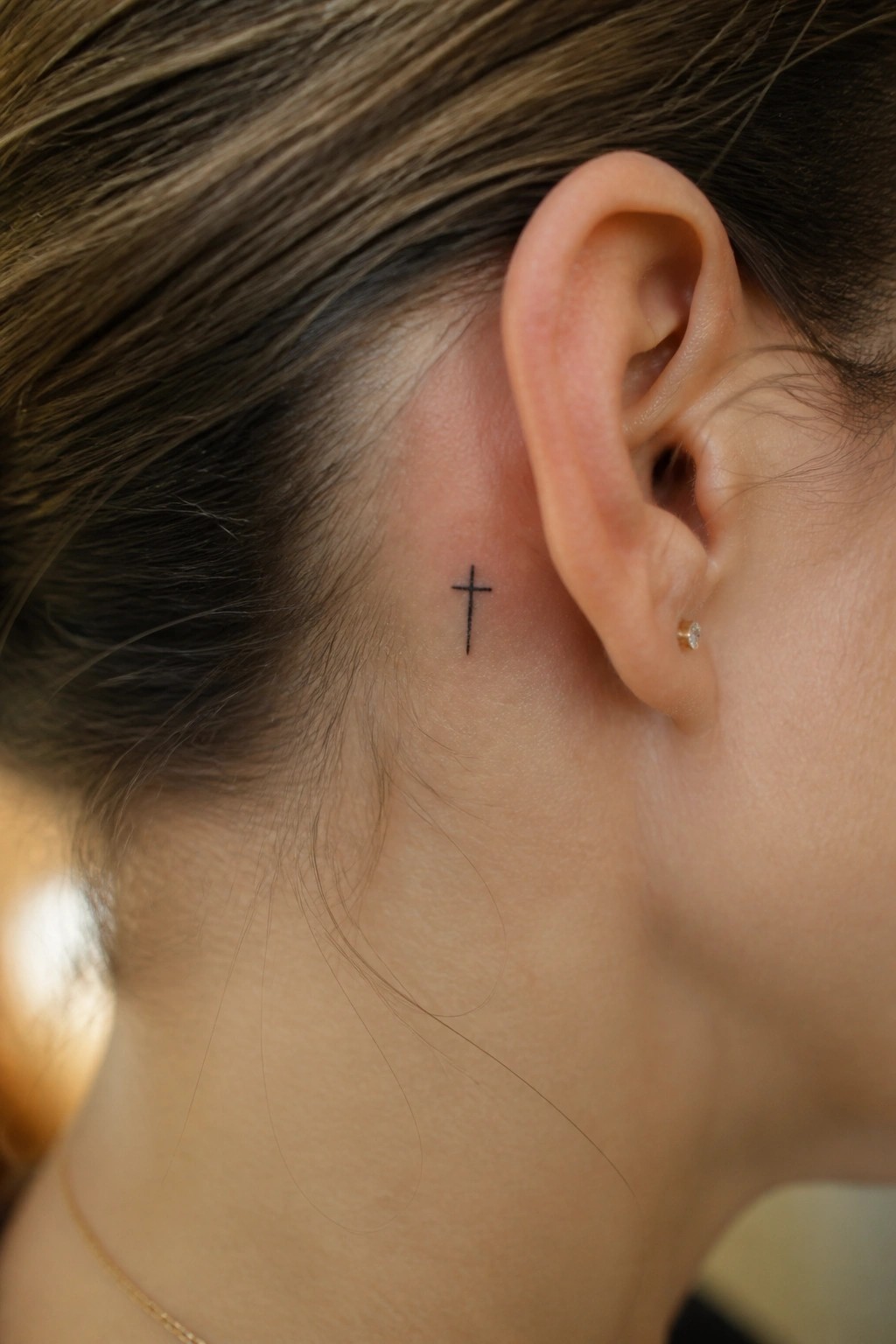 Small black cross tattoo behind ear with gold stud earring and necklace