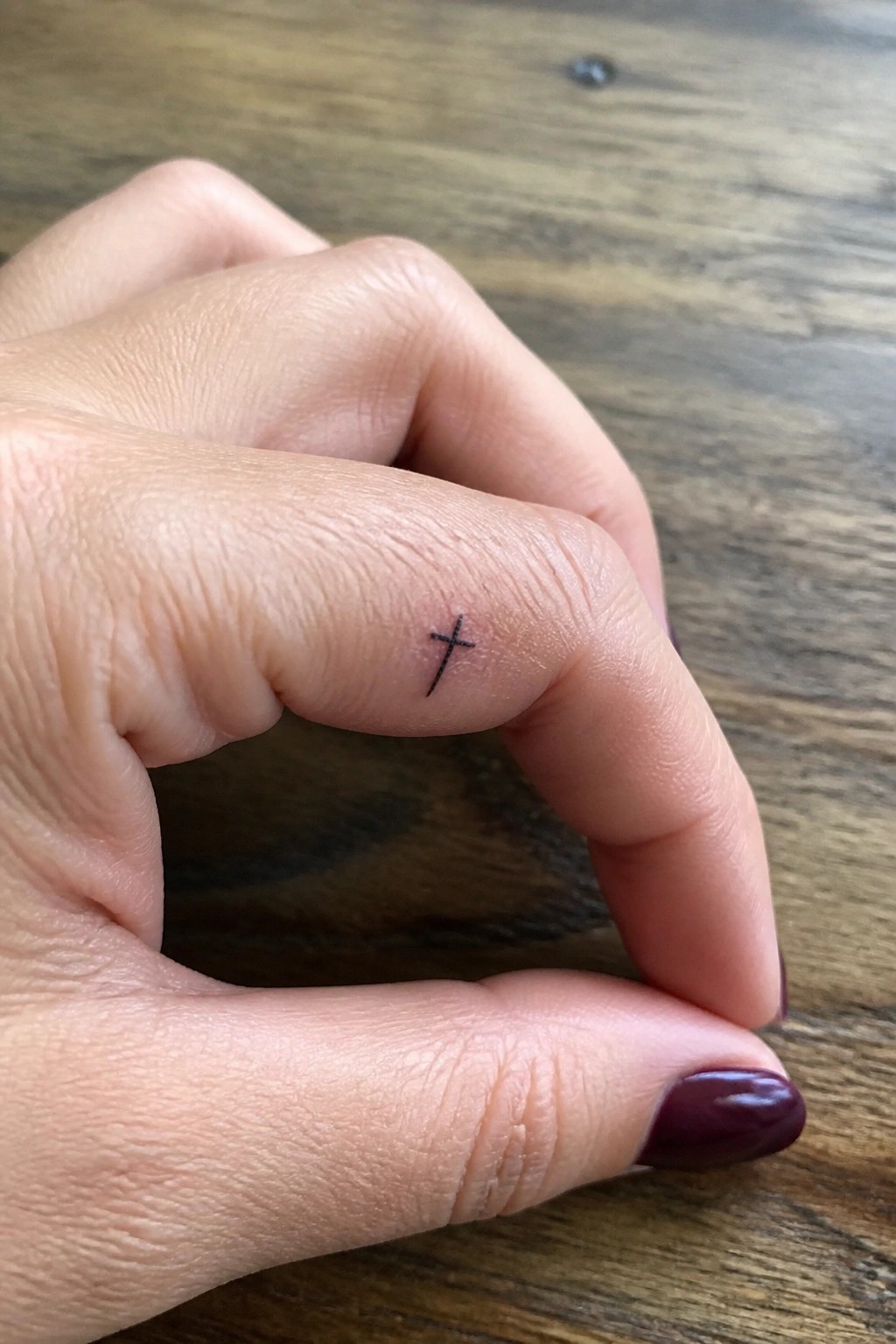 Hand with small black cross tattoo on index finger and dark maroon nails on wood