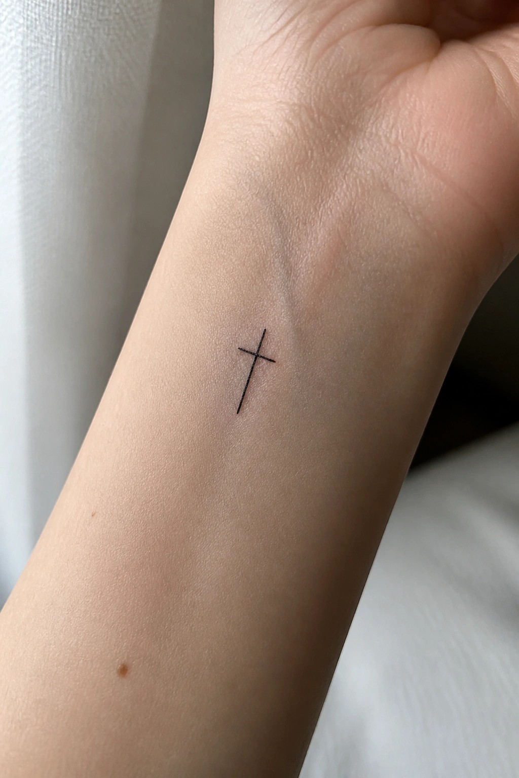 Small black cross tattoo on pale inner wrist beside tiny brown mole.