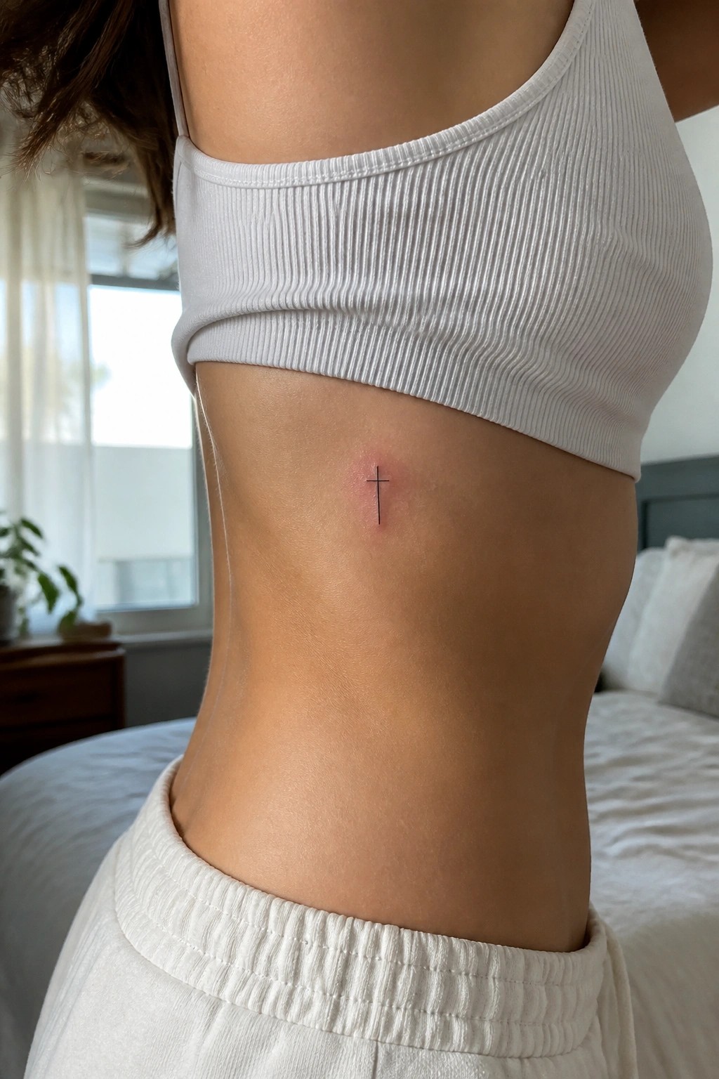 Small cross tattoo on ribcage below white ribbed crop top and sweatpants.