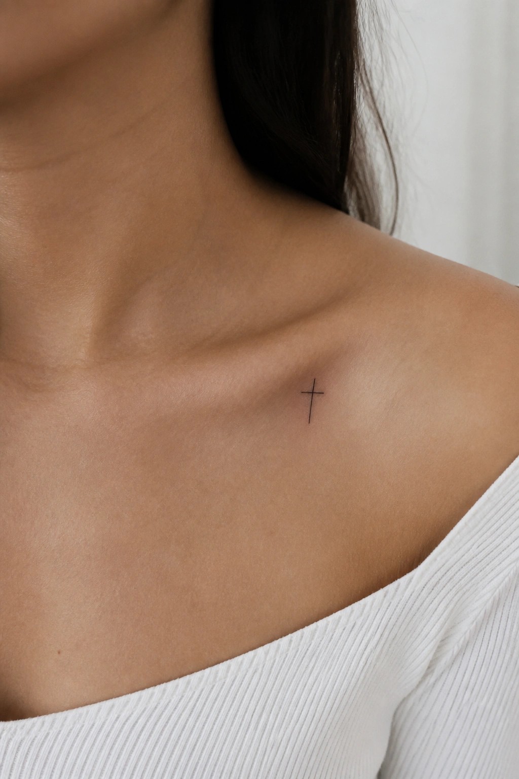 Small fine line cross tattoo placed on the upper chest near the collarbone.