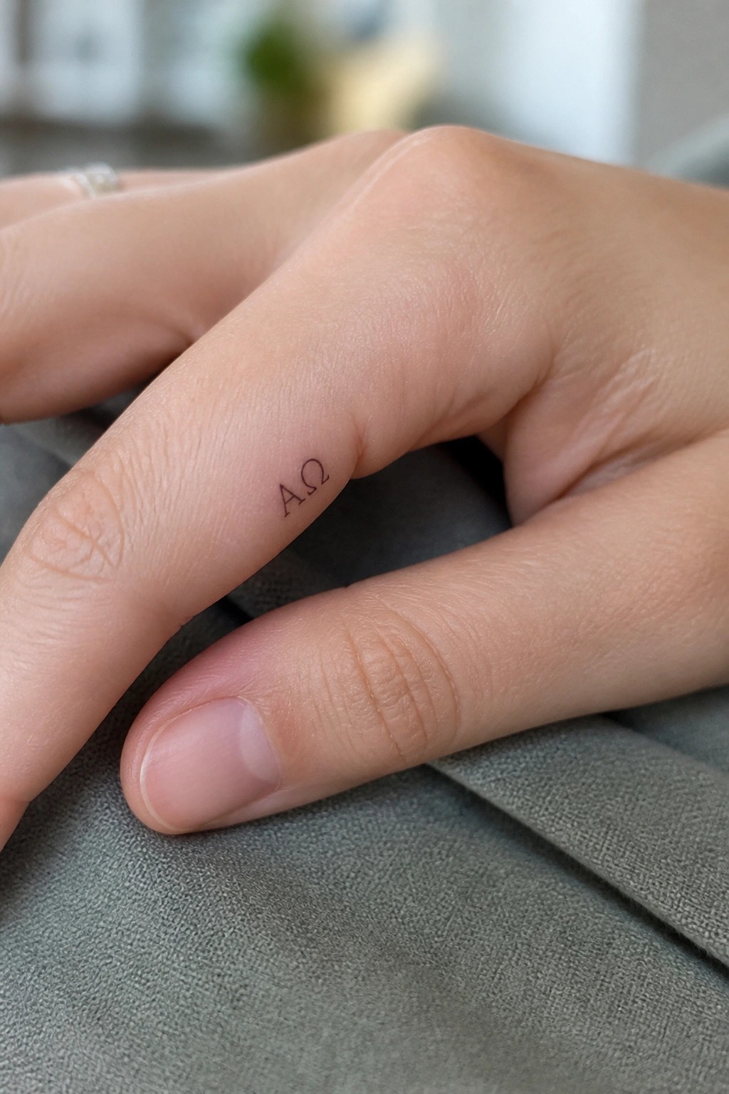 Hand with small AΩ tattoo on index finger resting on gray fabric
