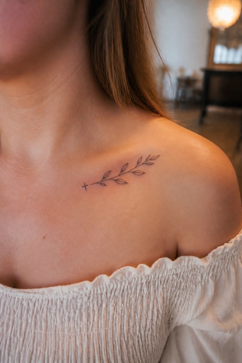 Woman's shoulder with small leafy branch and cross tattoo above off-shoulder white top
