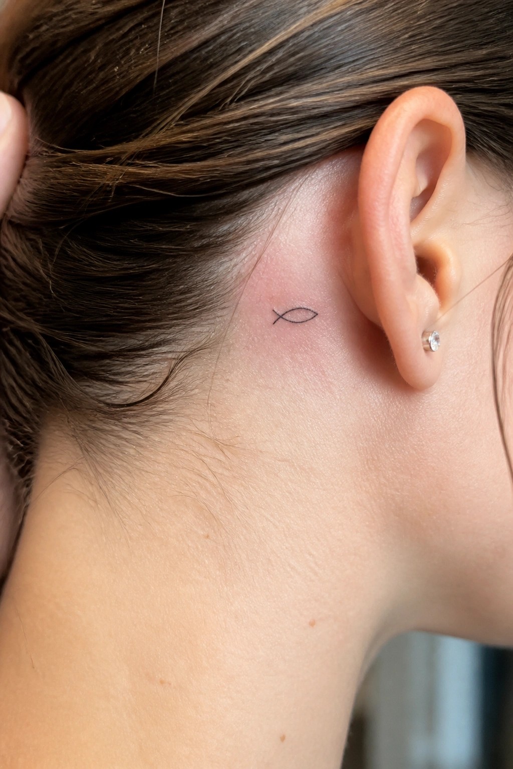 Small black outline fish tattoo placed behind the ear on a woman's head