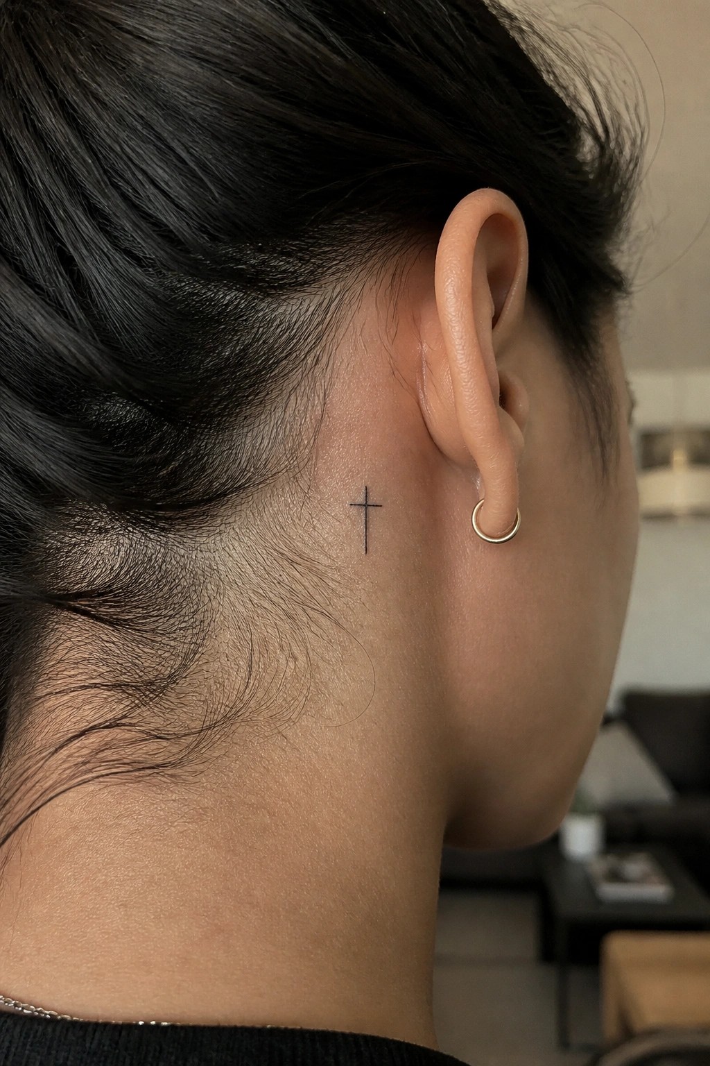 Small black cross tattoo placed behind the ear on the side of the head.