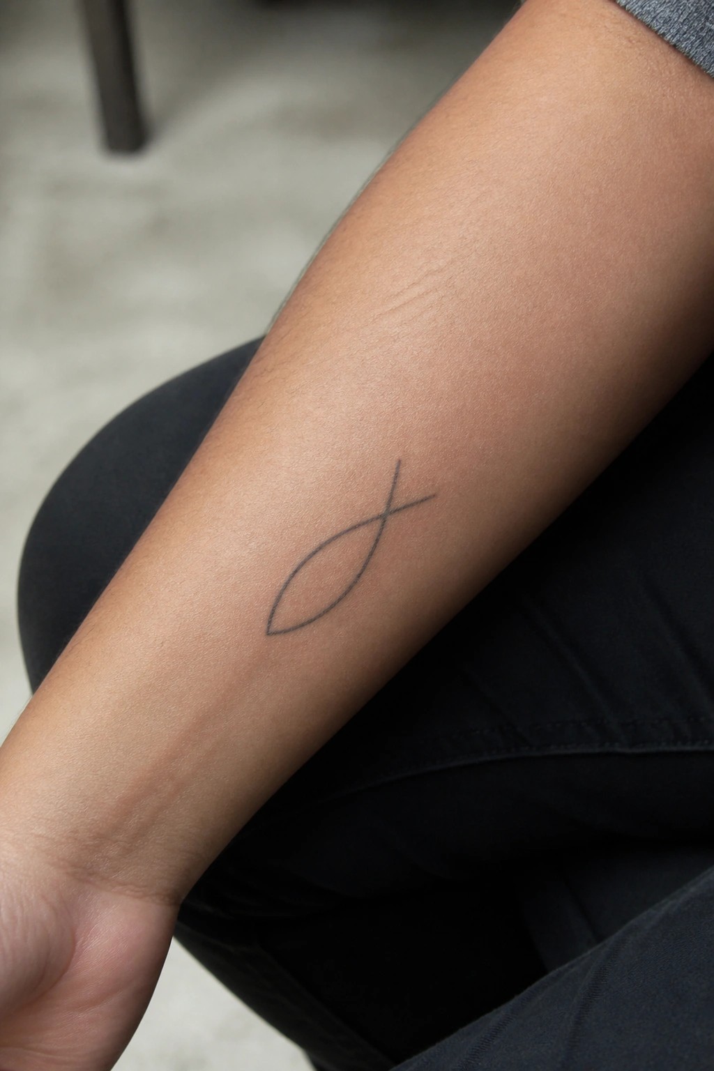 Person's forearm with minimalist black fish tattoo on light brown skin, black pants visible.