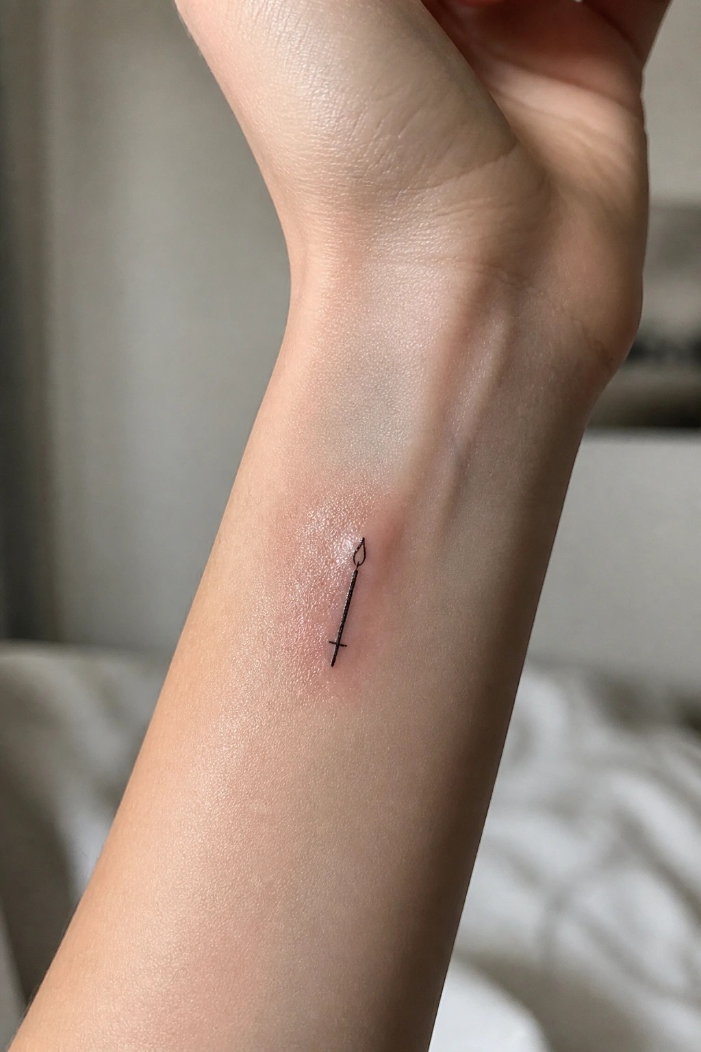 Small minimalist black line tattoo of a sword with flame tip and cross guard on the inner wrist.