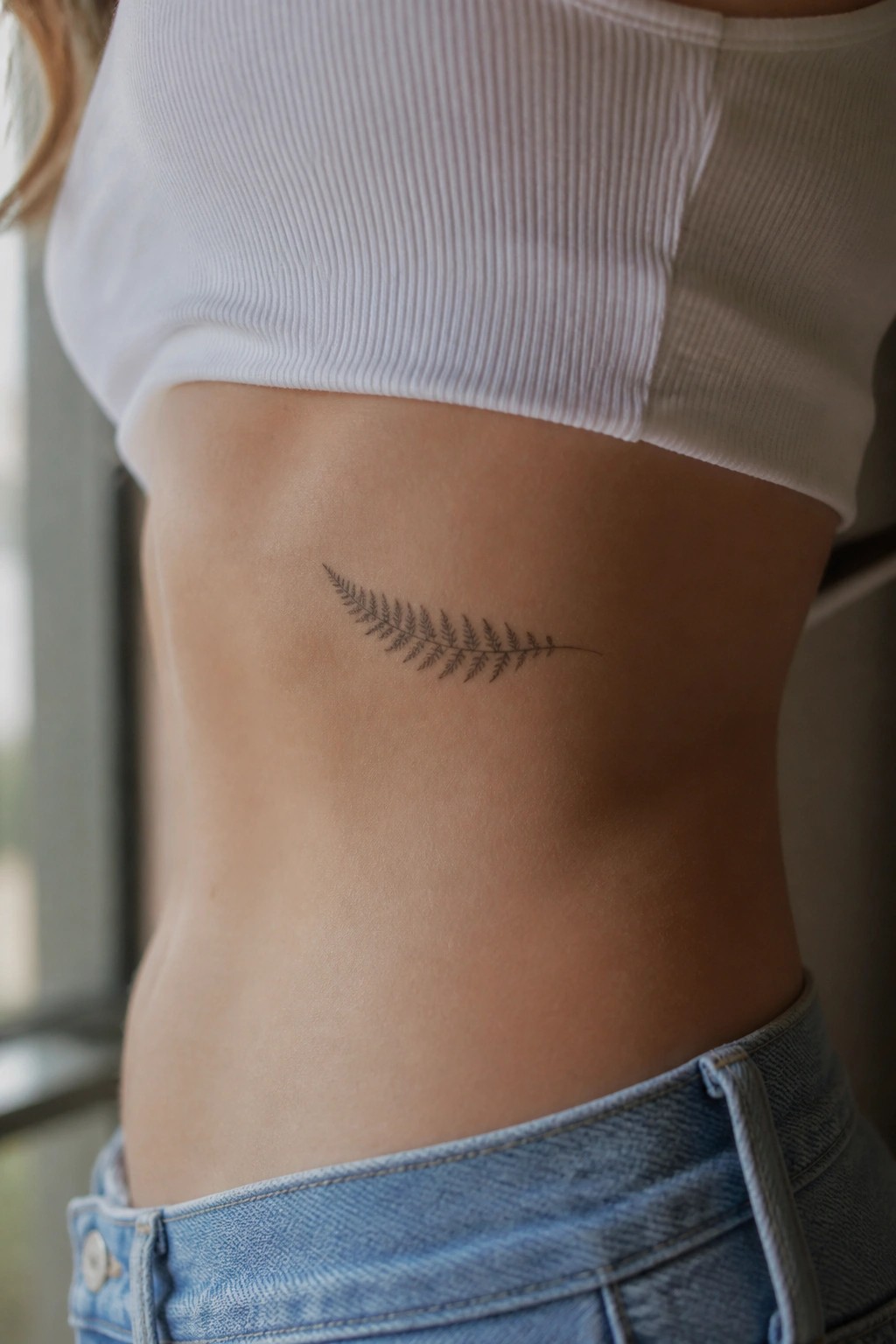 Fine line fern frond tattoo on the side of a woman's torso above the waistband of jeans.