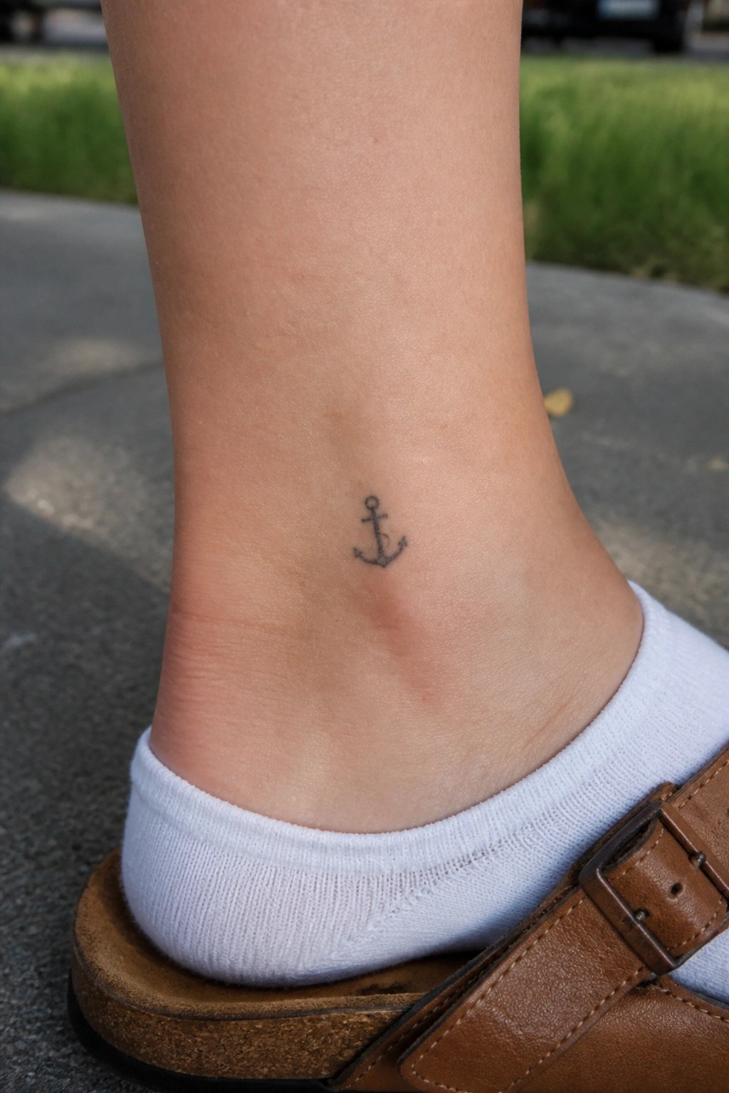 Small black anchor tattoo on the ankle above a white sock and brown sandal