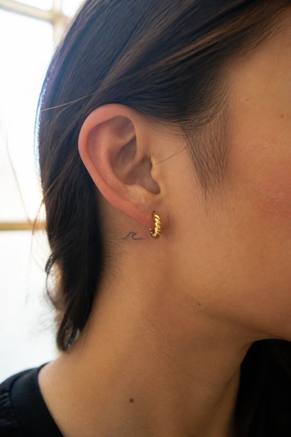 Close-up of ear with gold twisted hoop earring and small wave tattoo on neck