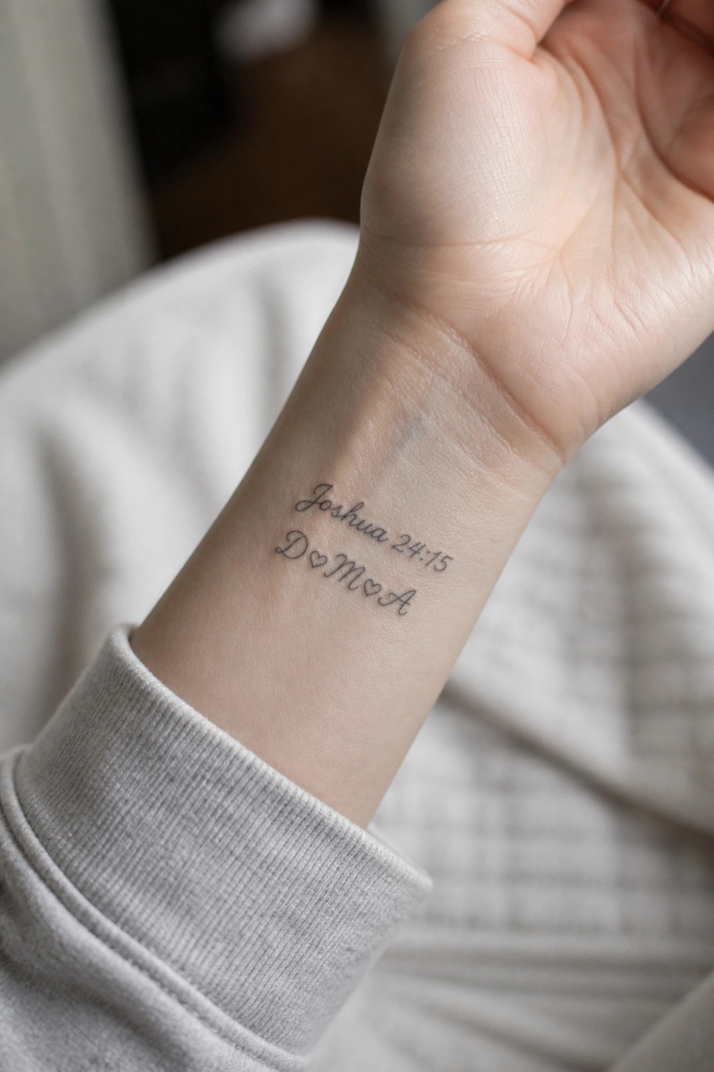 Wrist tattoo in black cursive reading Joshua 24:15 above D♡M♡A on gray sleeve.