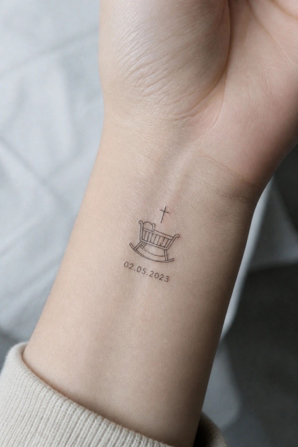 Wrist tattoo of rocking cradle with cross above and date 02.05.2023 below