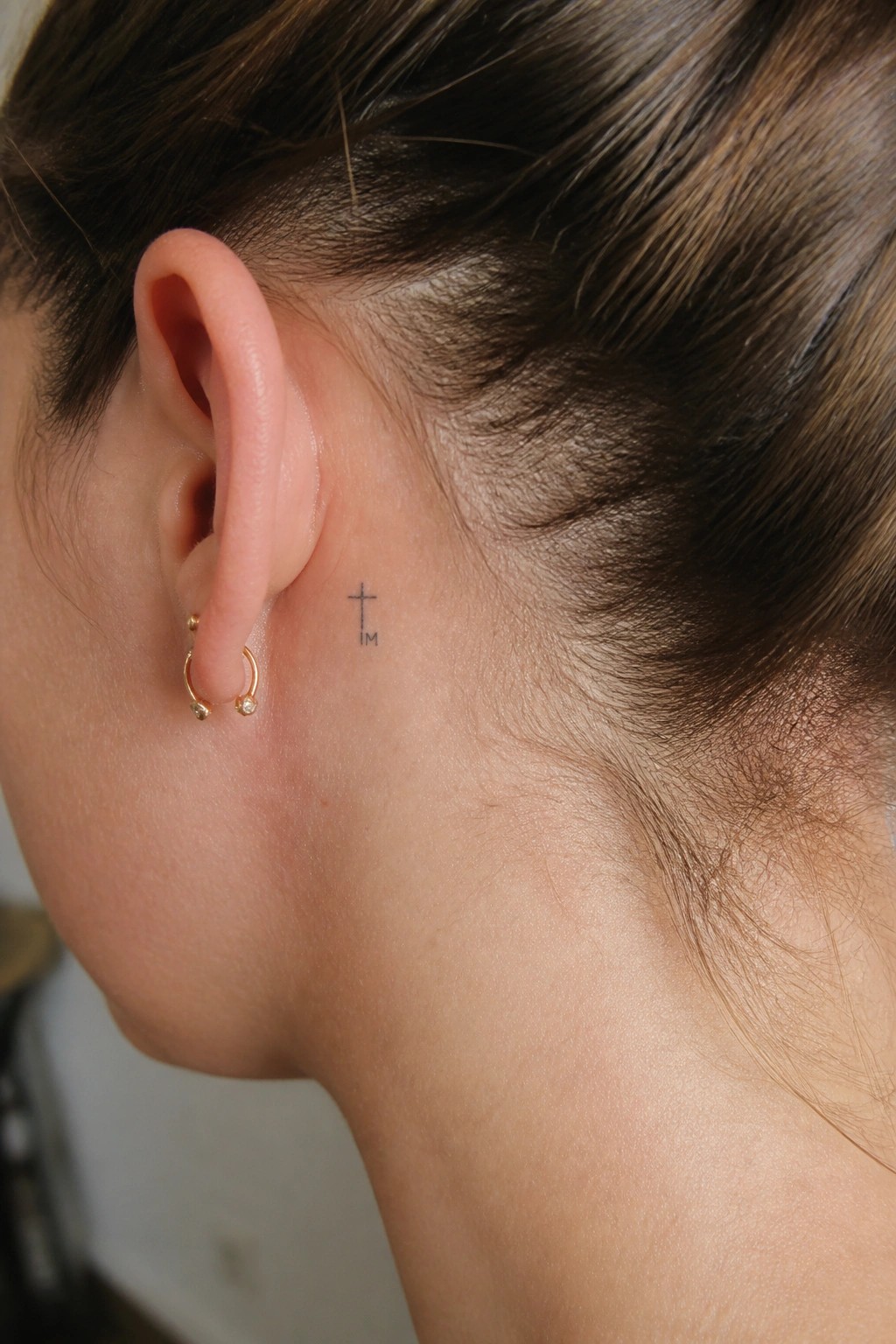 Ear with gold hoop earrings and small cross tattoo reading IM behind it