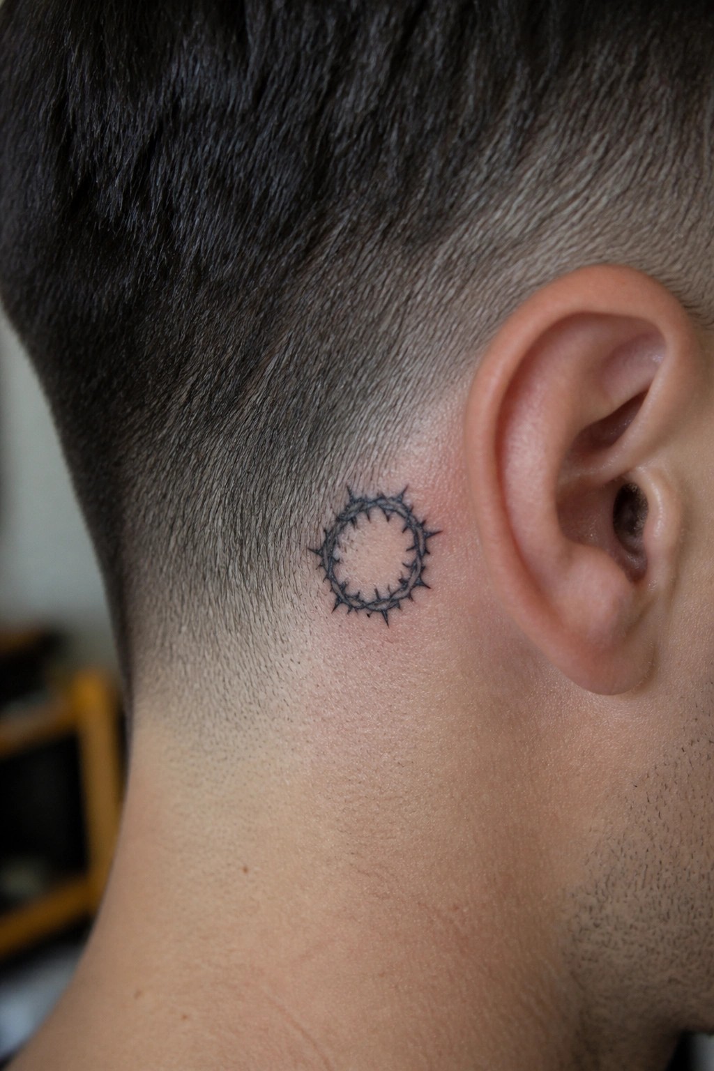 Small black crown of thorns tattoo in a circular shape on the side of the neck behind the ear.