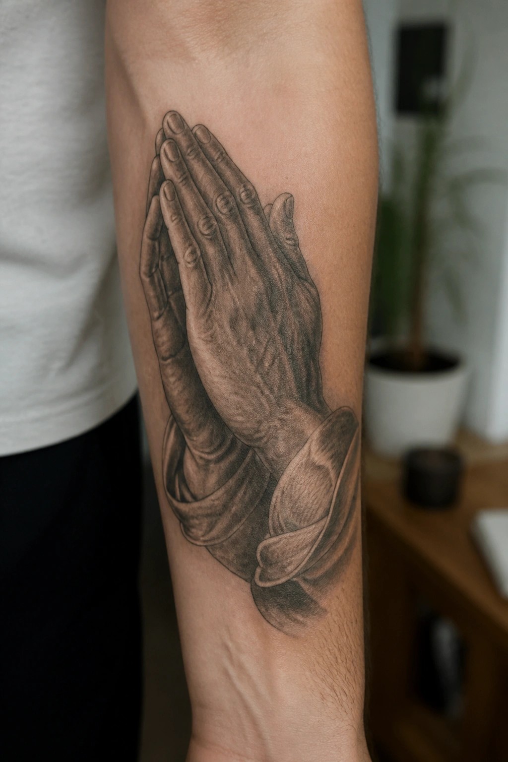 Realistic black-and-gray tattoo of praying hands on a forearm in casual attire.