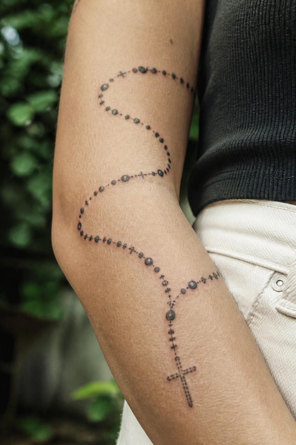 Rosary bead tattoo spirals along forearm ending in cross, dark shirt and white pants.