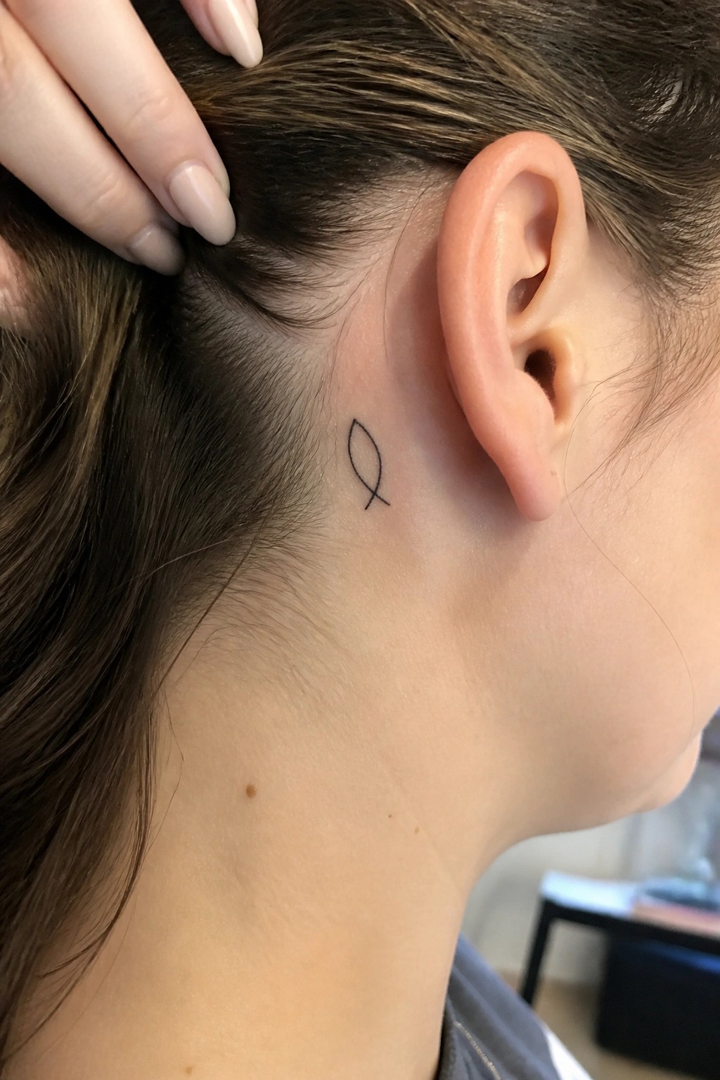 Small black outline of a Christian fish symbol tattoo placed behind the ear on the side of the head