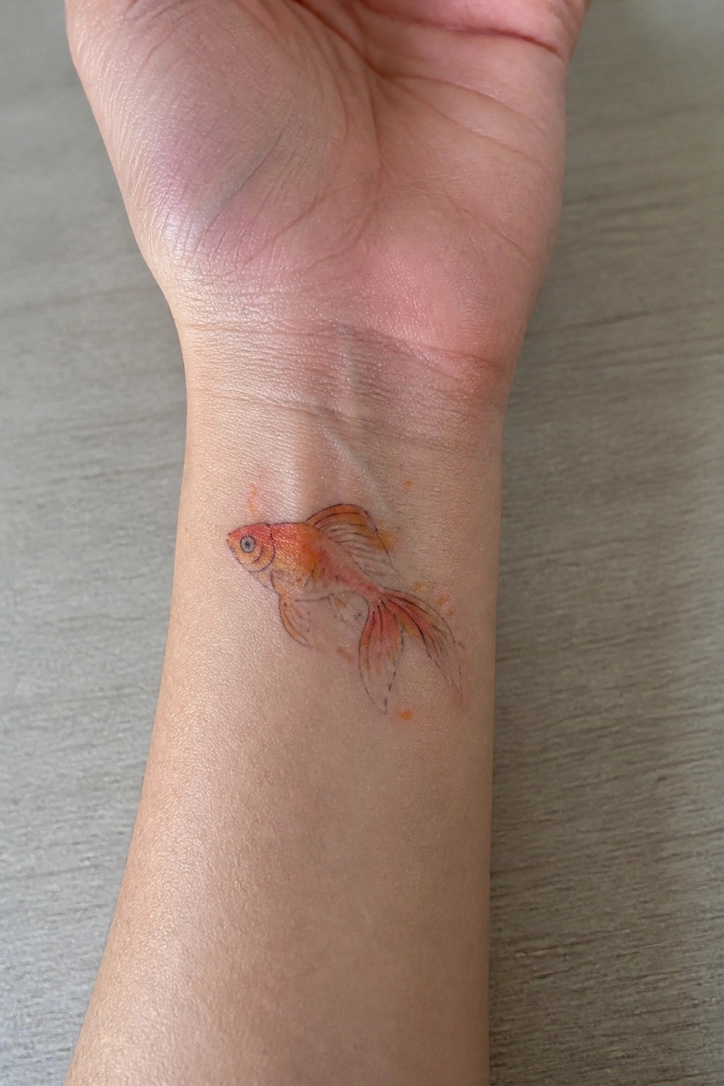 Small orange goldfish tattoo with soft watercolor shading placed on the inner wrist