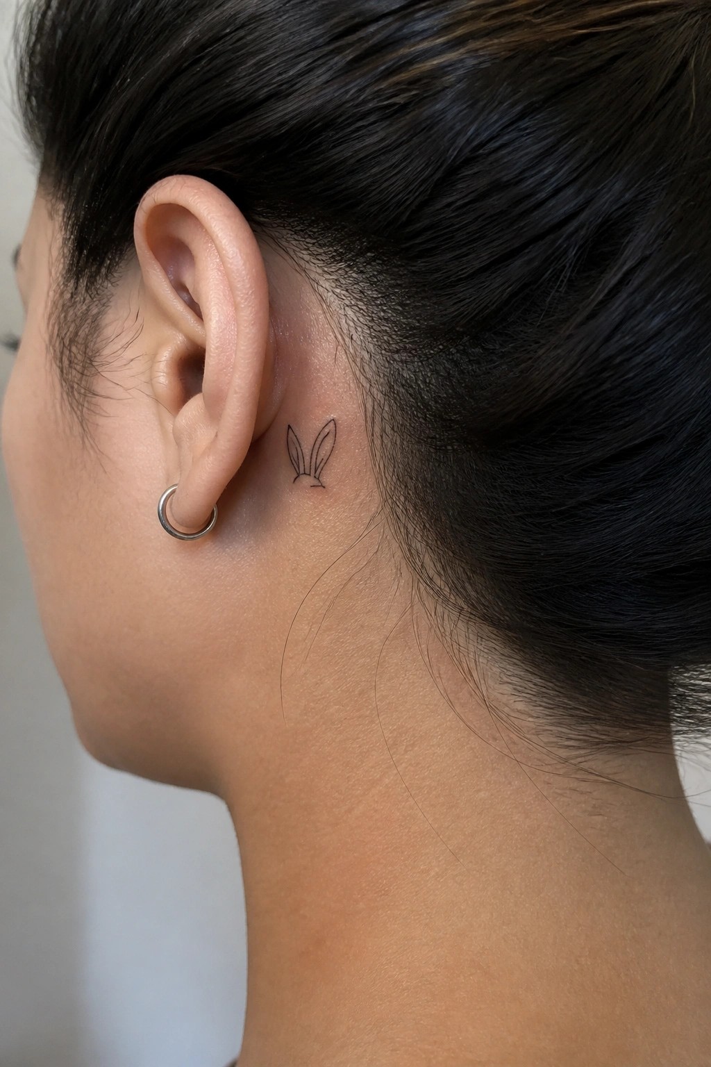 Close-up of ear with silver hoop earring and small black bunny ears tattoo behind it.