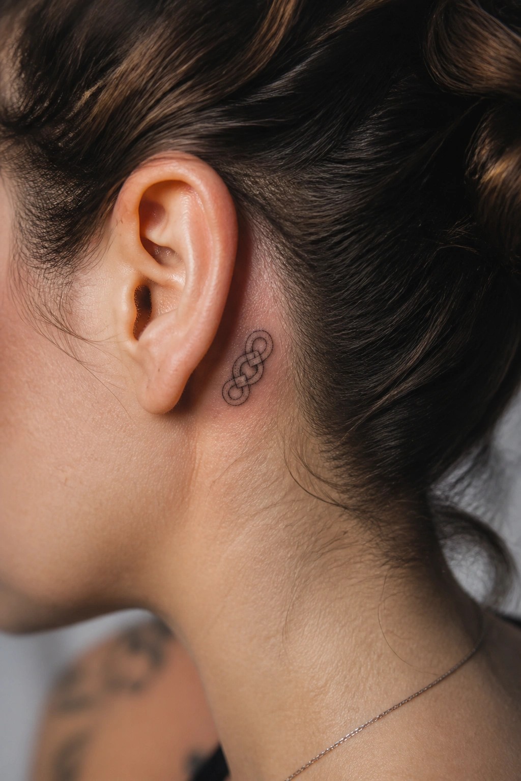 Woman's ear with small black intertwined loop tattoo and thin necklace.