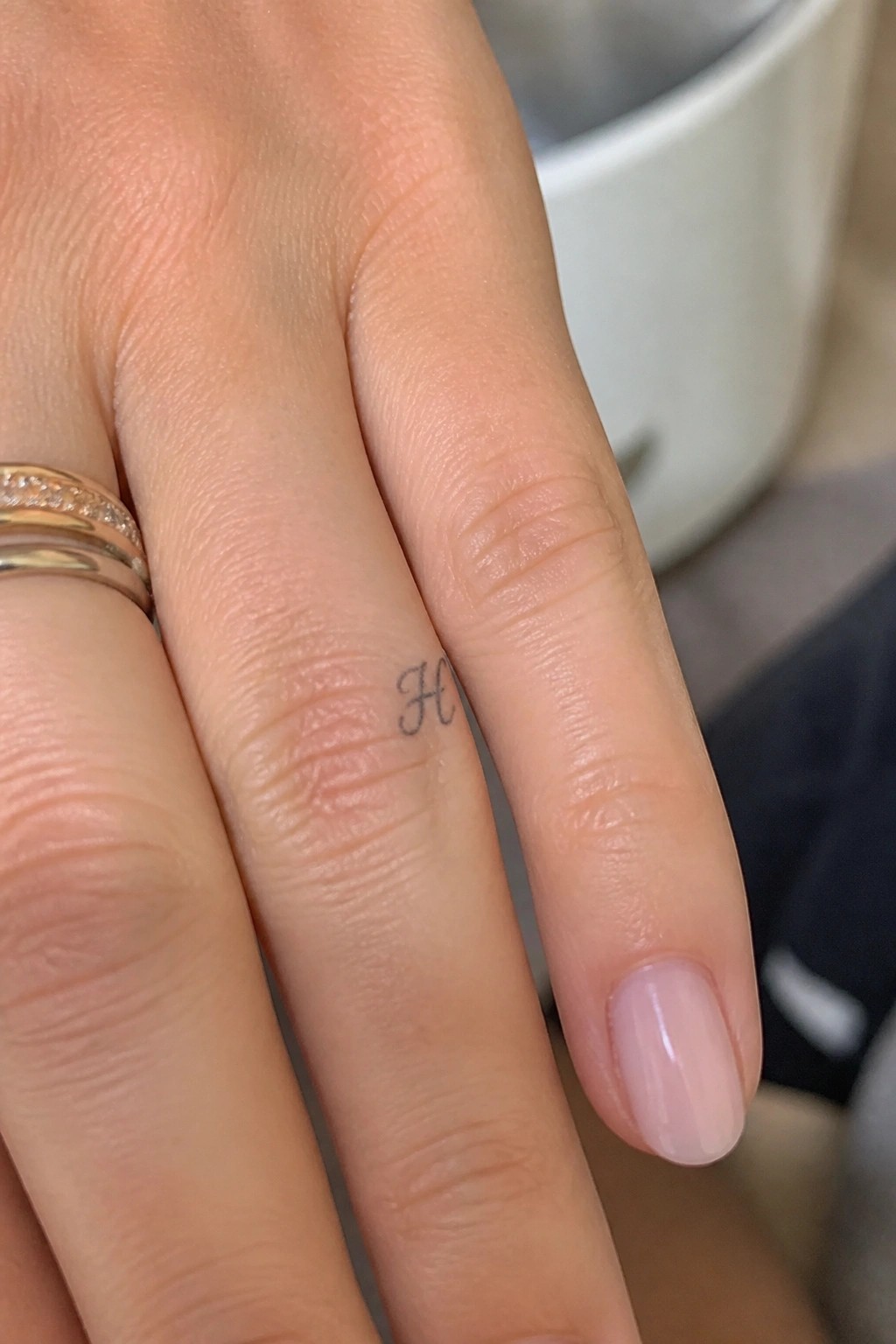 Fingers with light pink nail polish, cursive H tattoo, and two gold rings