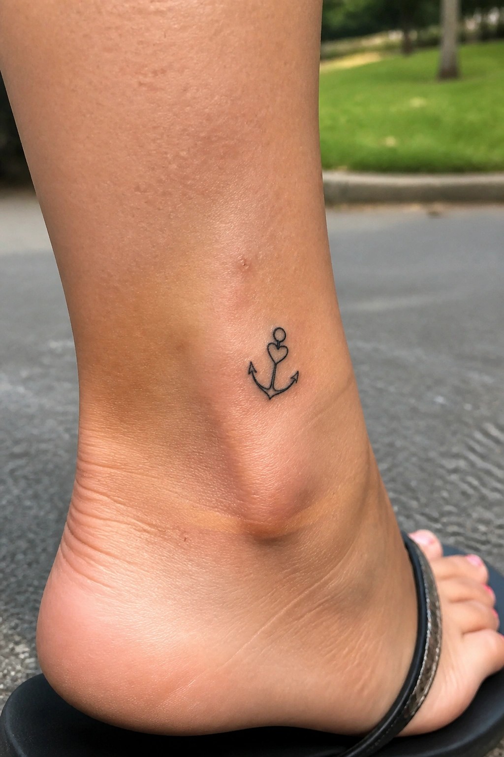 Small black line anchor tattoo with a heart on the shank, placed on the outer ankle.