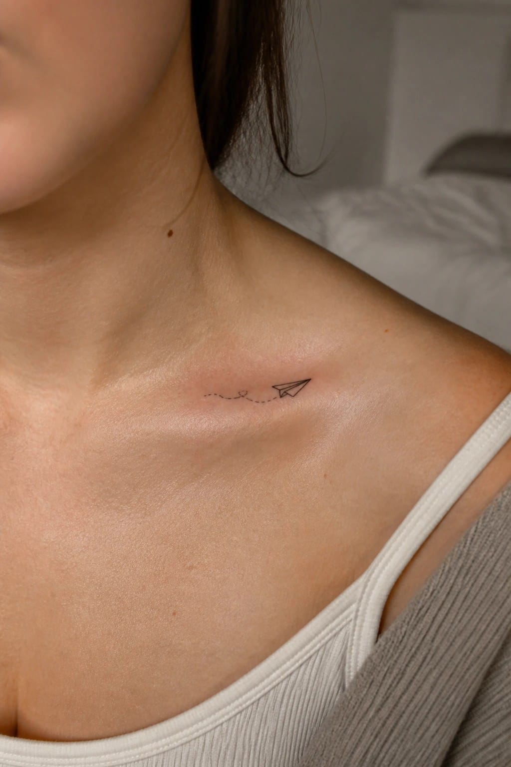 Paper airplane tattoo with dotted line and heart on collarbone above white tank top