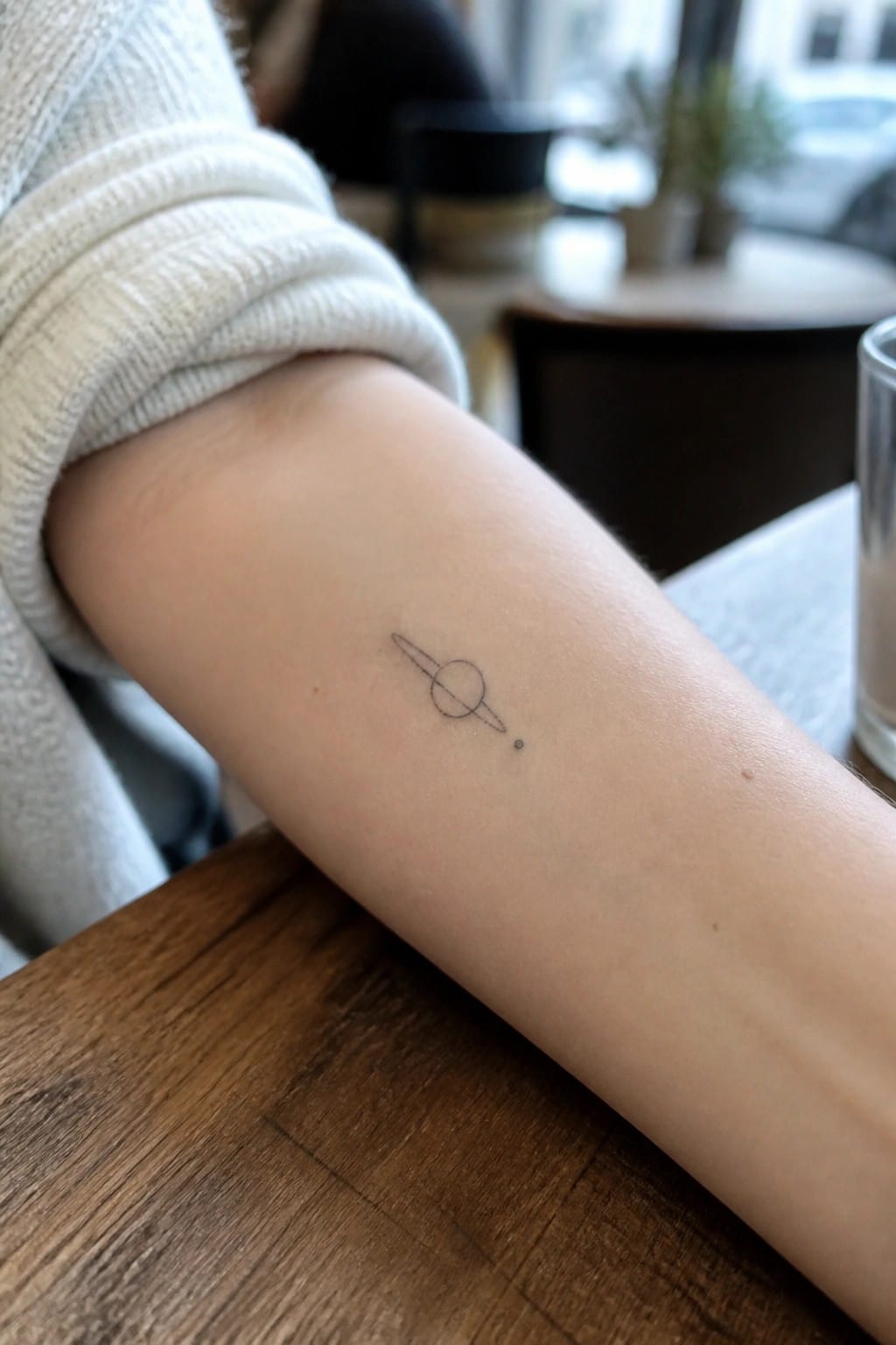 White knit sleeve rolled up on arm with small Saturn tattoo on wooden table