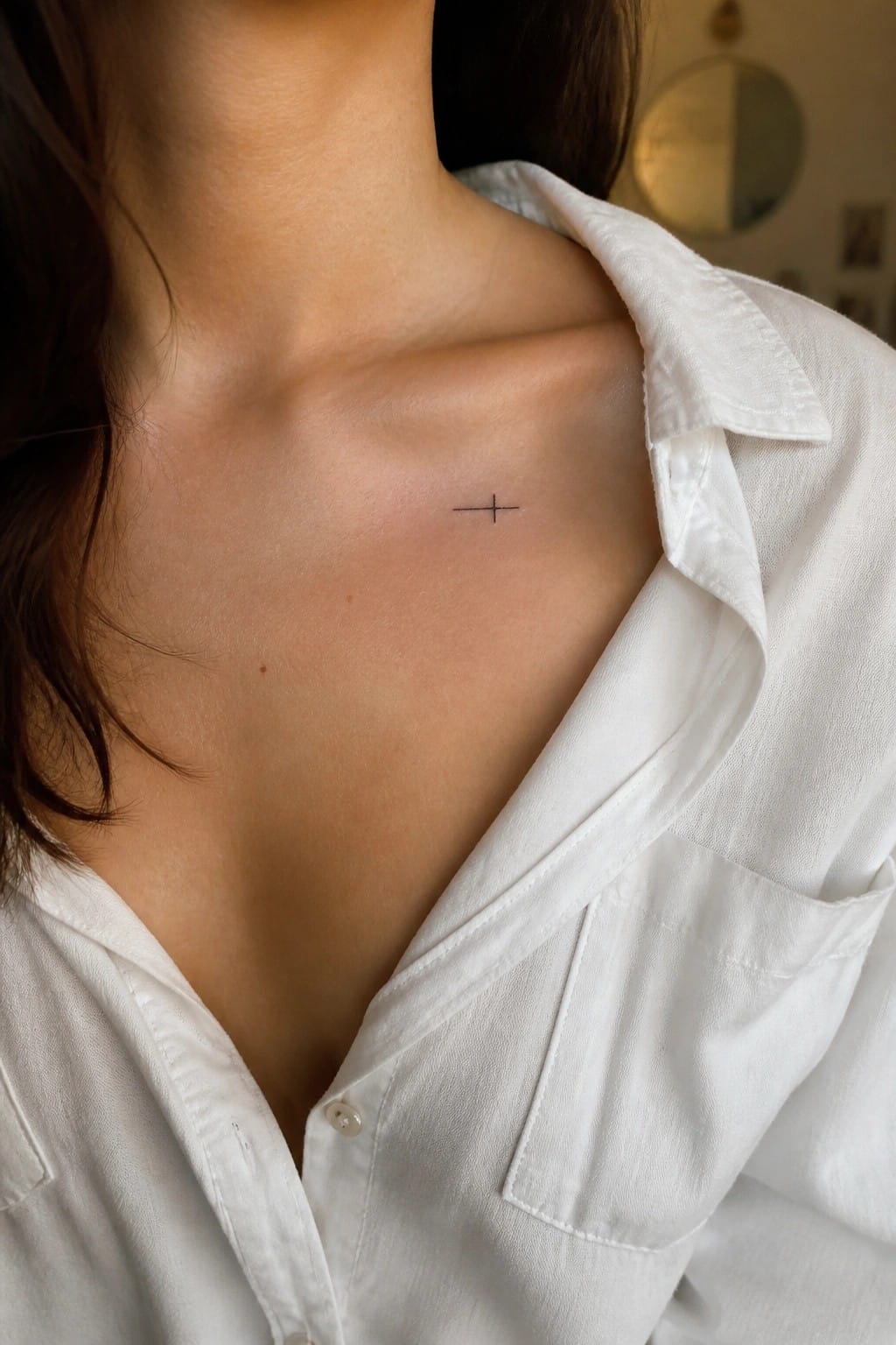 Close up of a small fine line cross tattoo on a woman's upper chest near the collarbone, partially visible under an open white shirt collar.