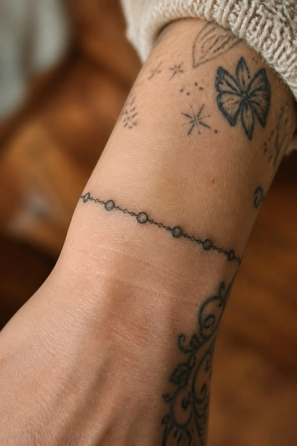 Wrist with black tattoos of dotted chain, flower, stars, and swirls under cream knit sleeve.
