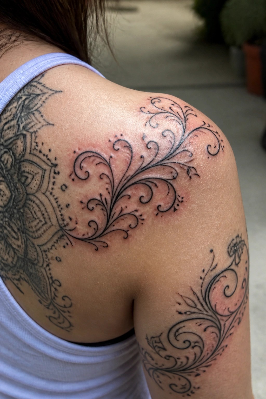 Black swirling filigree tattoo on a woman's shoulder next to a larger mandala, showing fresh ink with some redness.