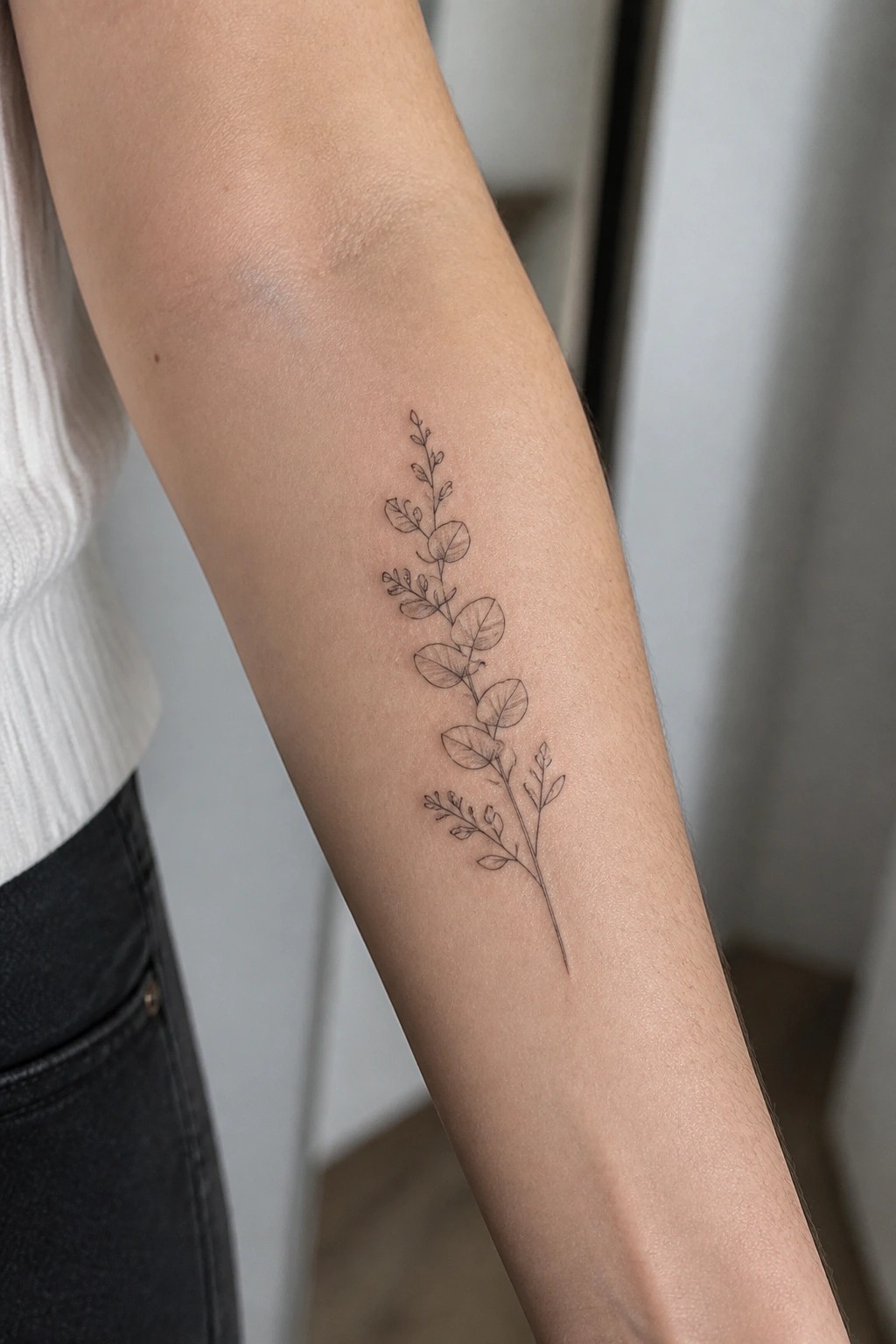Person's forearm with fine-line black eucalyptus tattoo, white top and dark jeans visible.