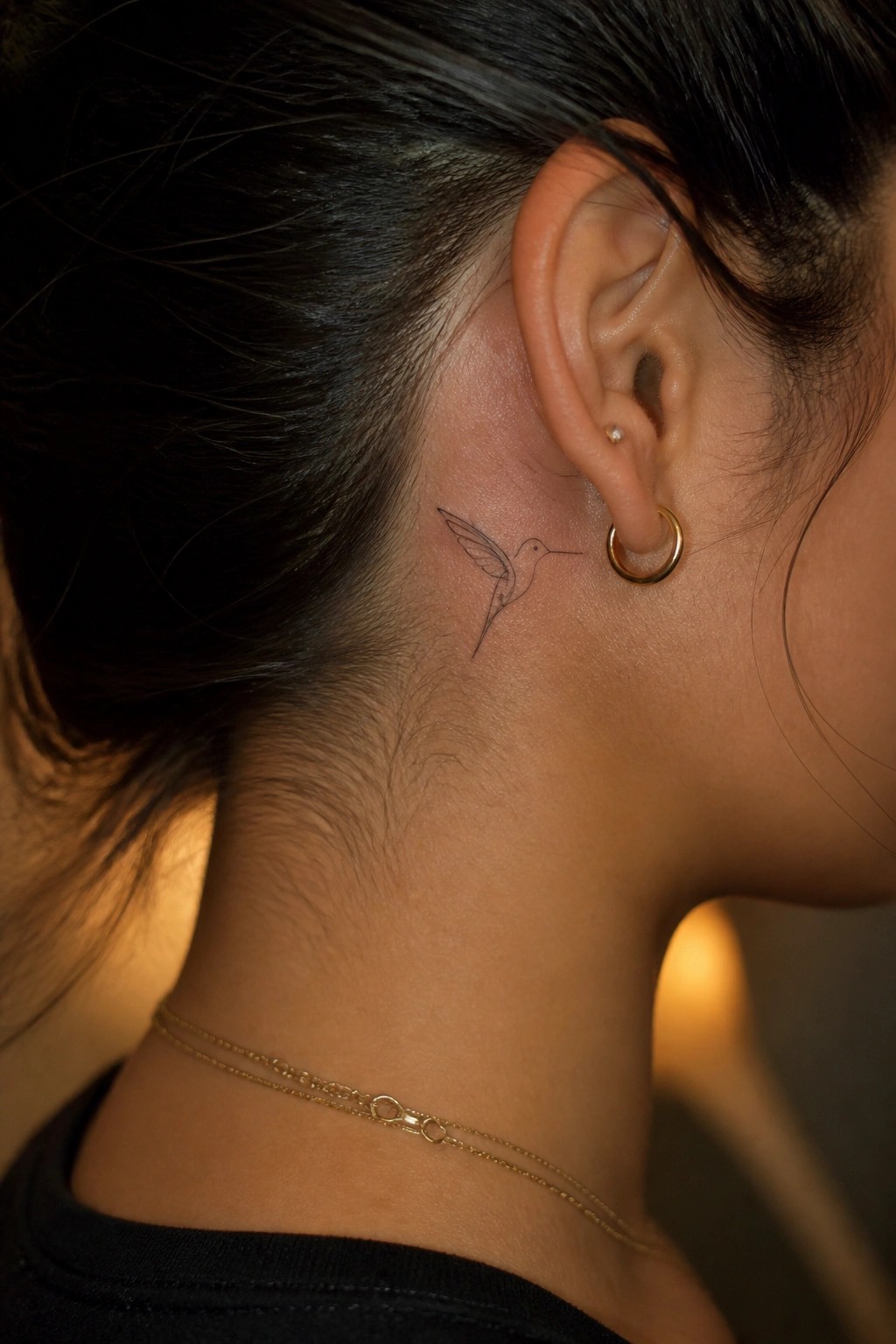 Hummingbird tattoo behind ear on neck with gold hoop earring and necklaces