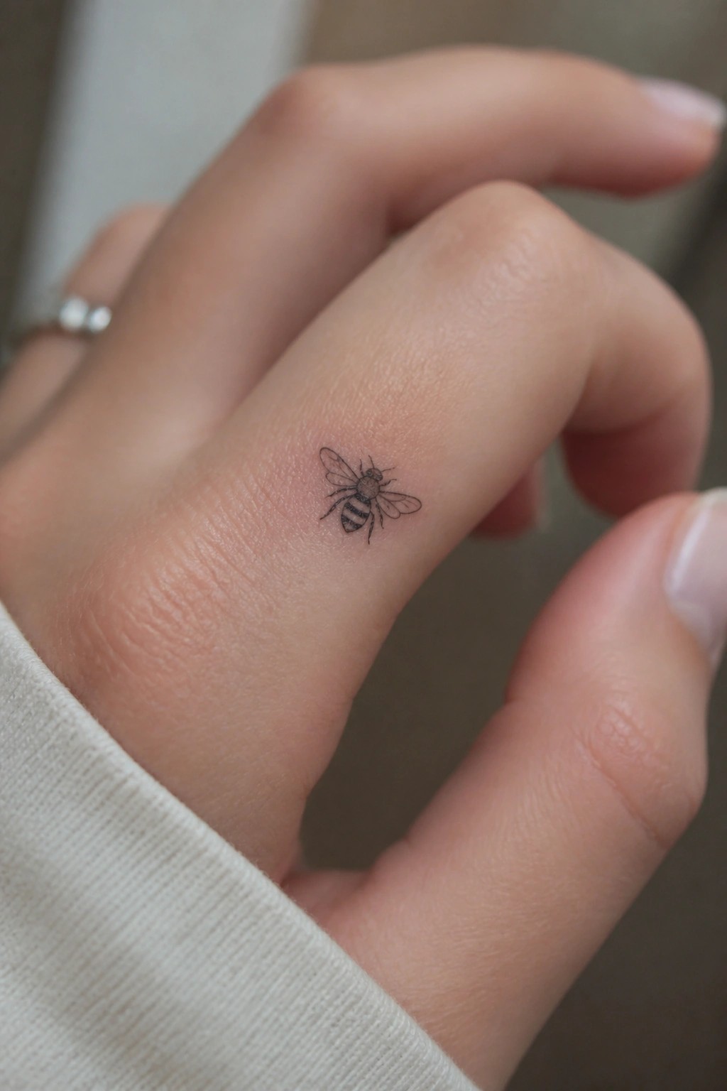 Light-skinned hand with white sleeve, small bee tattoo, and silver ring on finger.