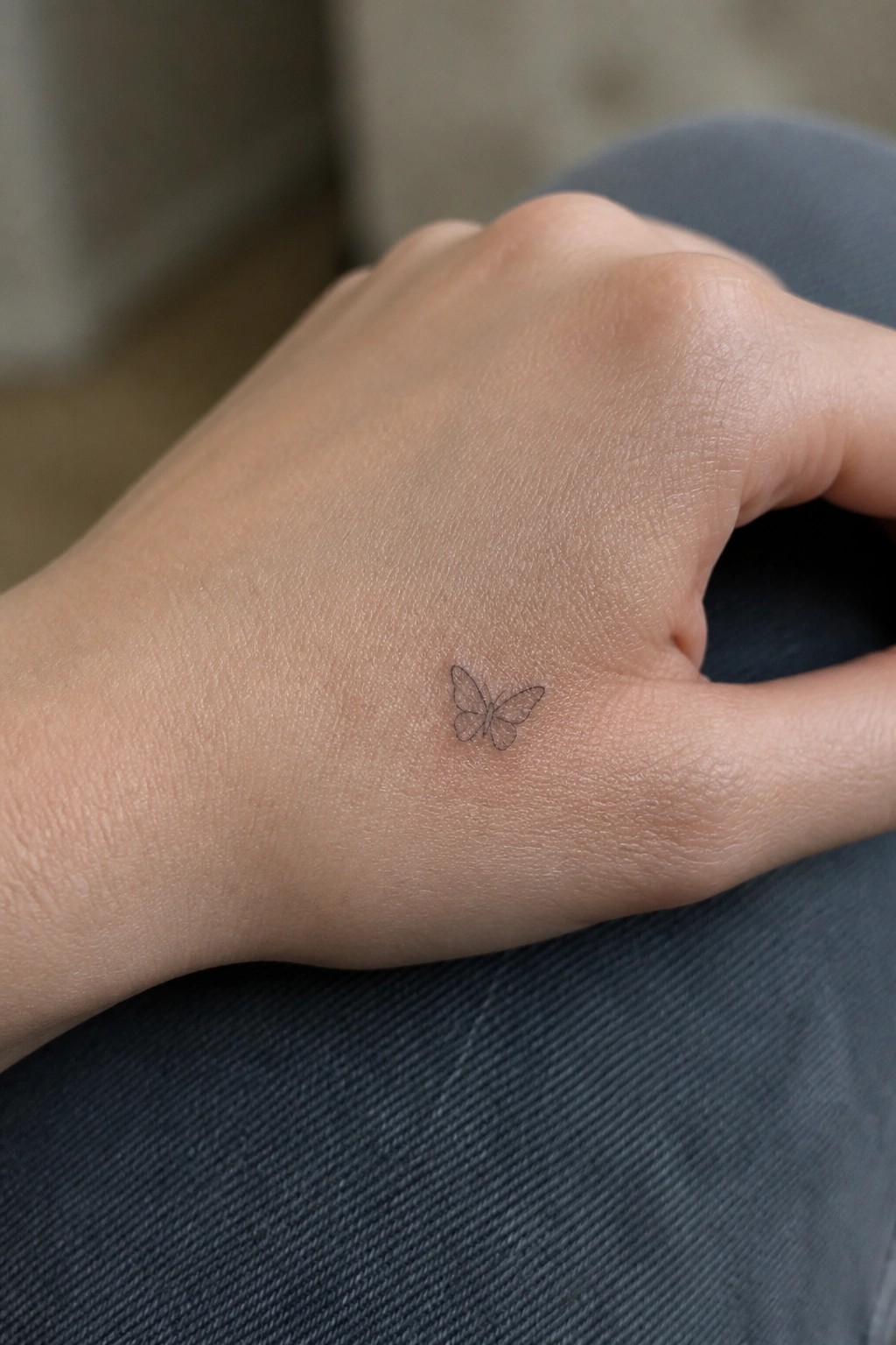 Small black outline butterfly tattoo on the side of a hand in minimal line style
