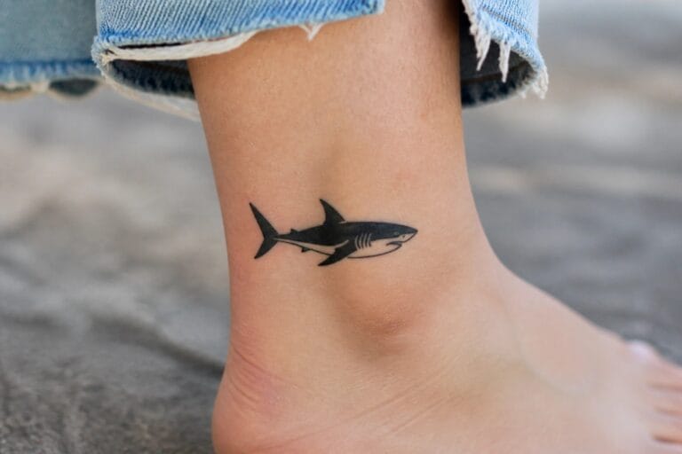 23 Bold Small Shark Tattoo Ideas Inspired by Ocean Adventures Black shark tattoo on ankle beneath rolled gray denim pants outdoors