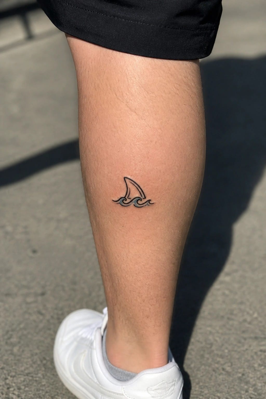 Small shark fin and wave tattoo on leg above white sneaker and black shorts
