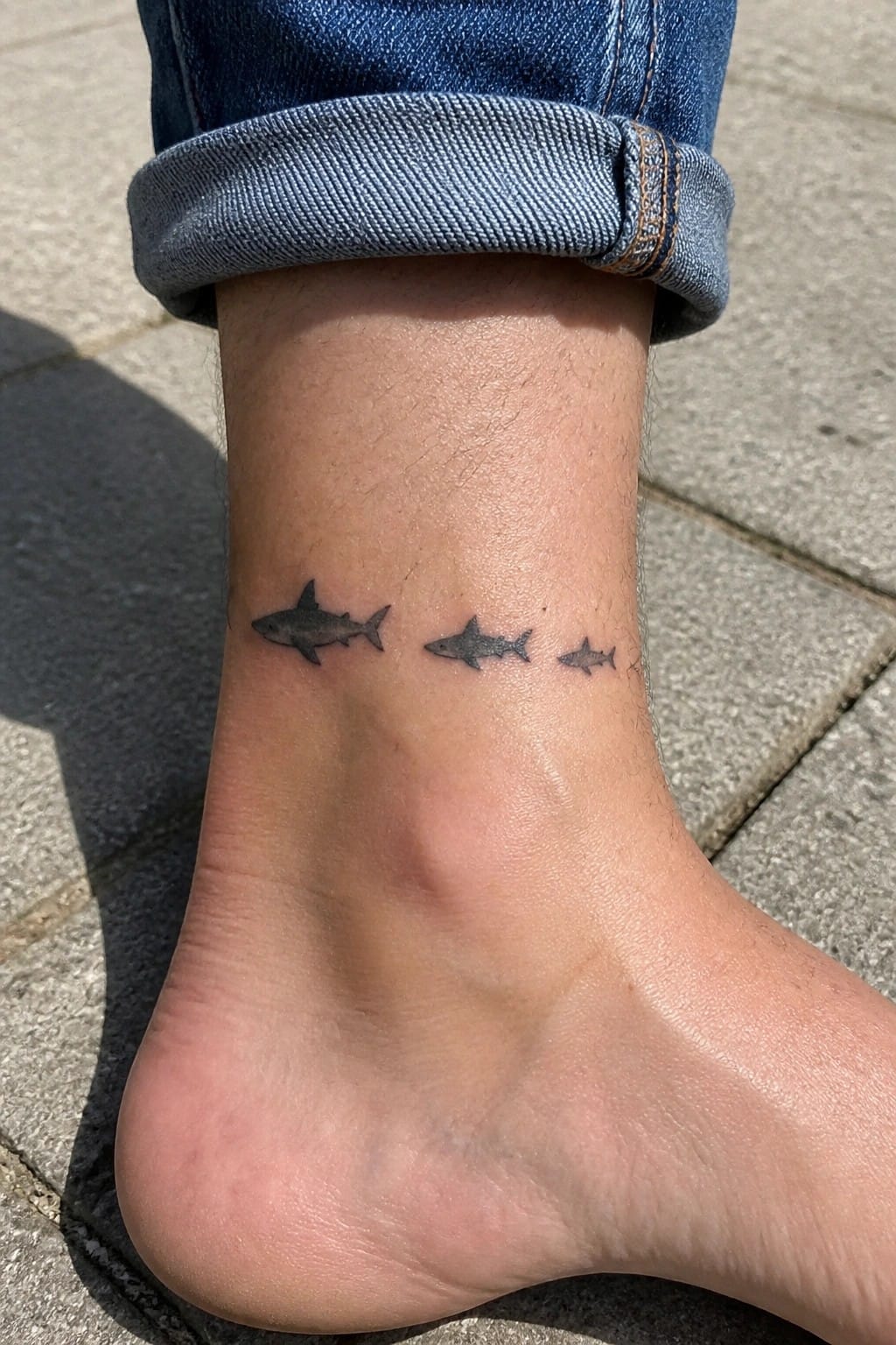 Three small black shark tattoos on ankle below rolled blue jeans cuff.