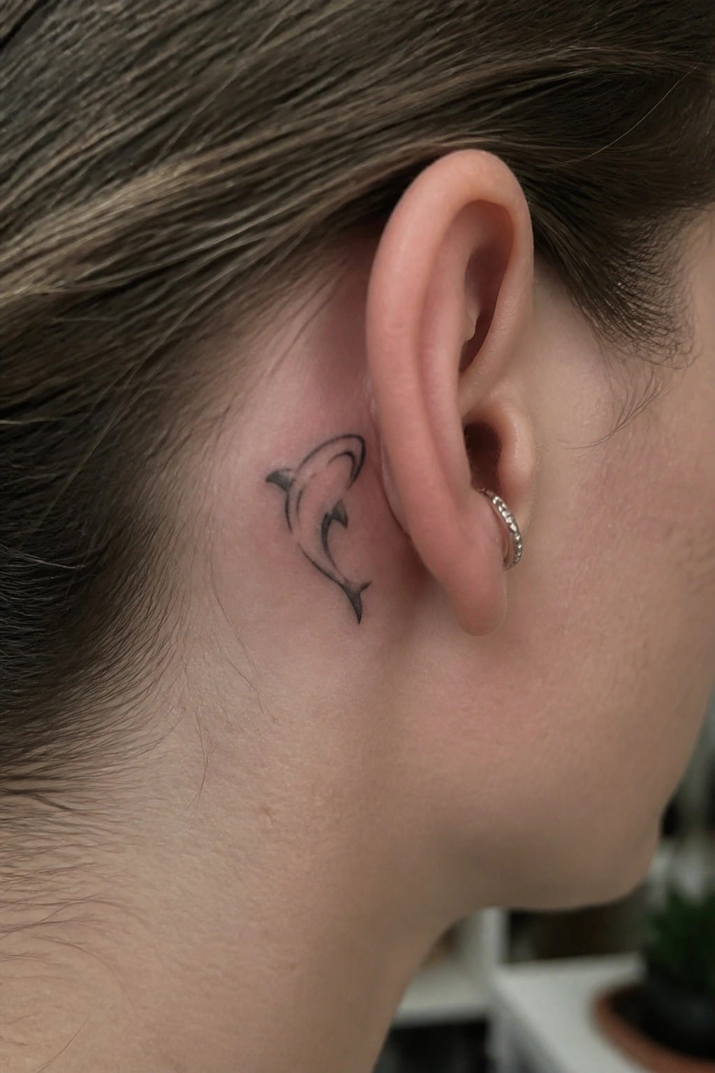 Small black outline dolphin tattoo placed behind the ear on a person's head.