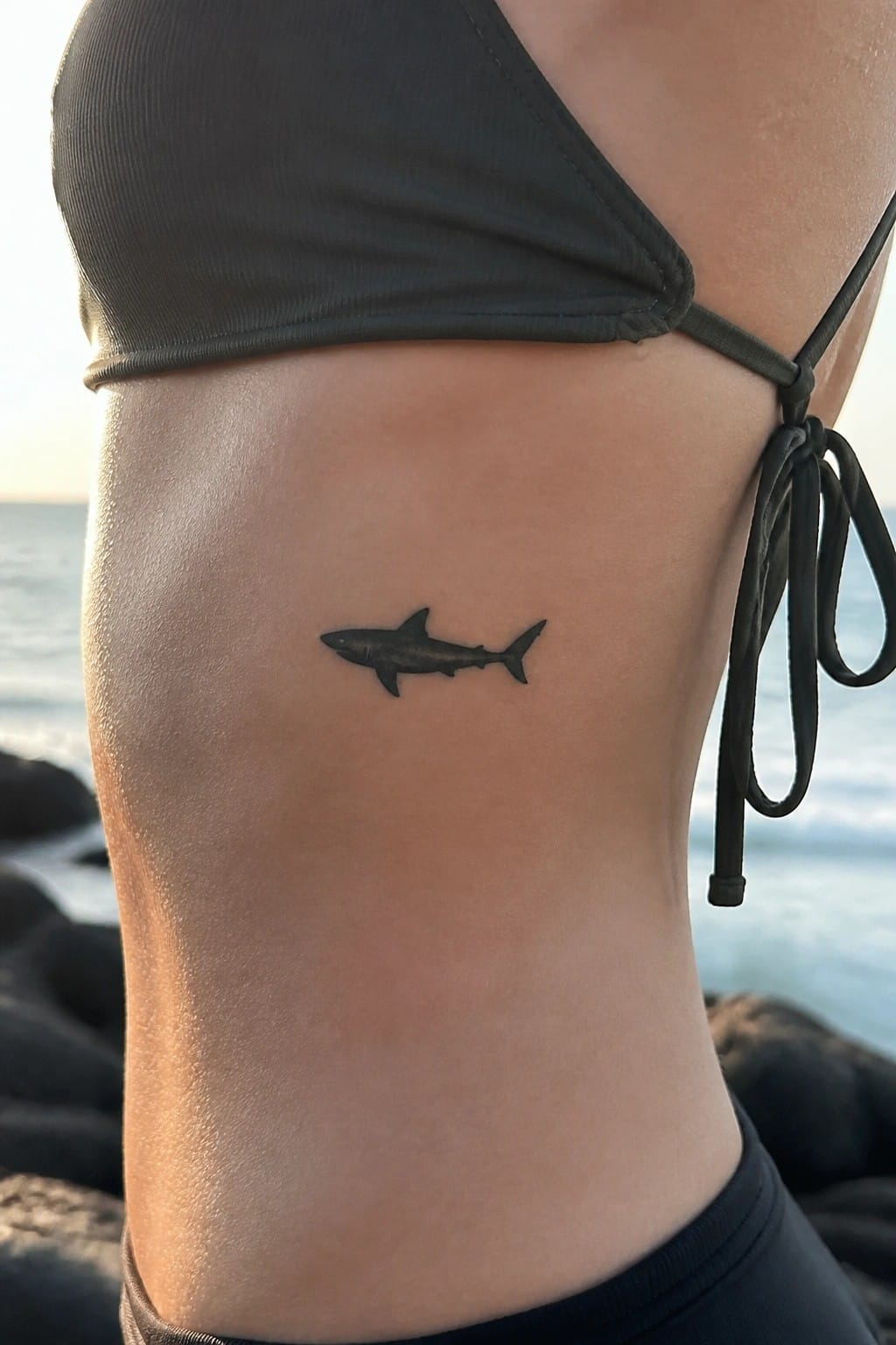 Black bikini top with small black shark tattoo on ribs by ocean at sunset