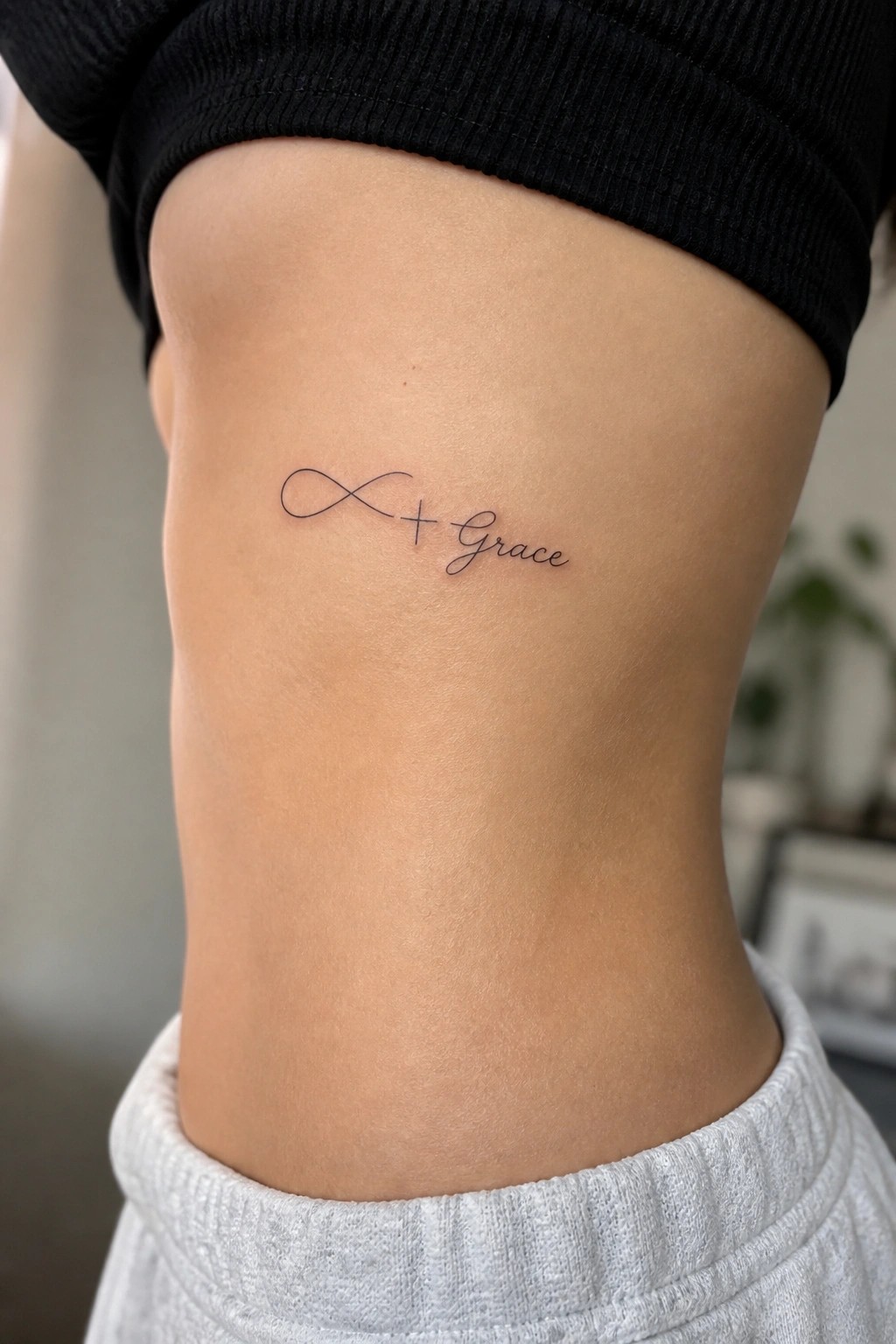 Infinity symbol plus Grace tattoo on skin below black top and gray pants