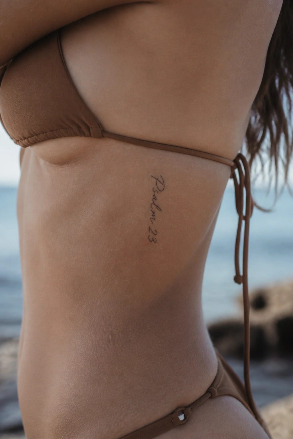Woman in brown bikini with Psalm 23 tattoo on side, ocean backdrop.