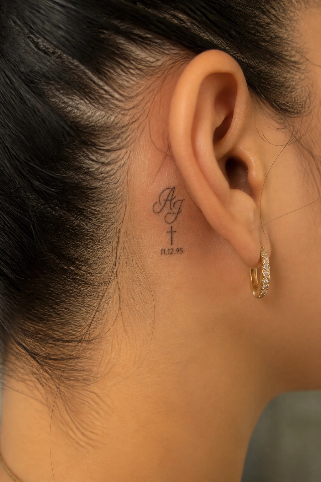 Close-up of ear with cursive 'Ag' tattoo, cross, date 11.12.95, and jeweled gold hoop earring