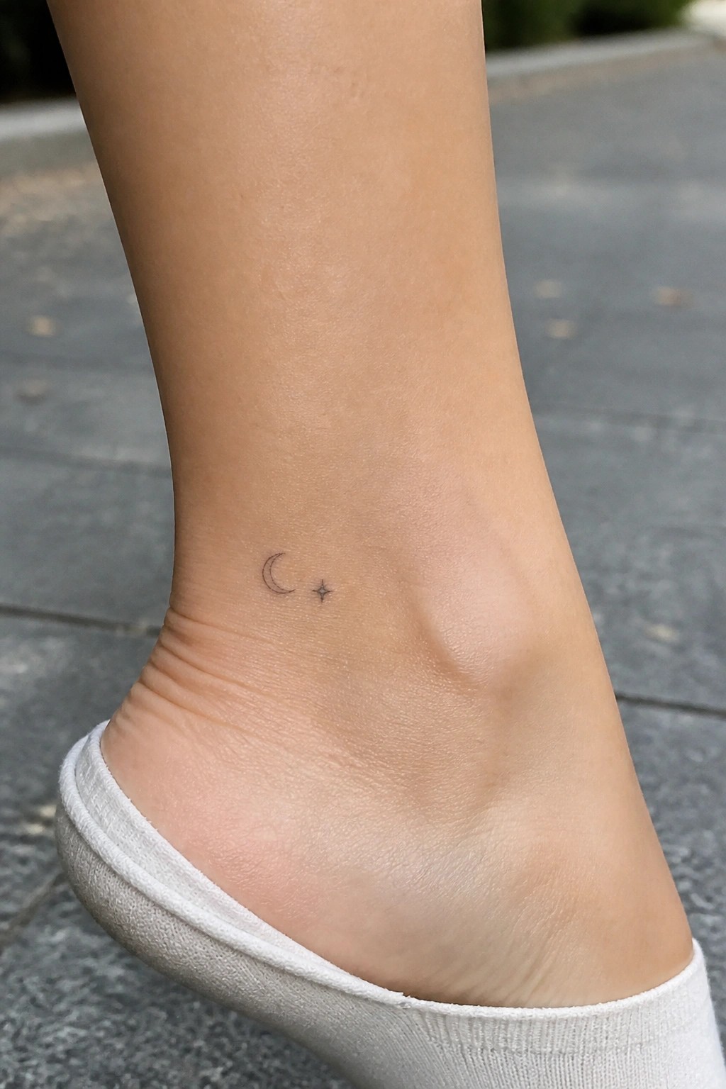 Minimalist crescent moon and star tattoo on light-skinned ankle above white sock on pavement.