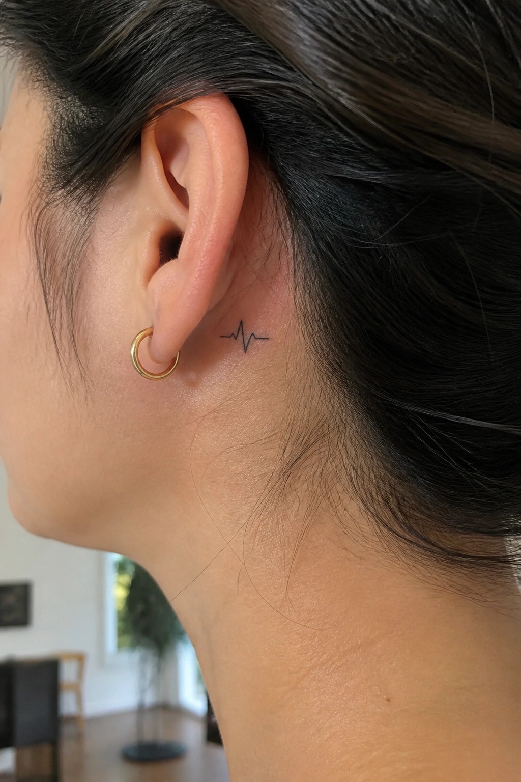 Small black heartbeat line tattoo placed behind a woman's ear next to a gold hoop earring.