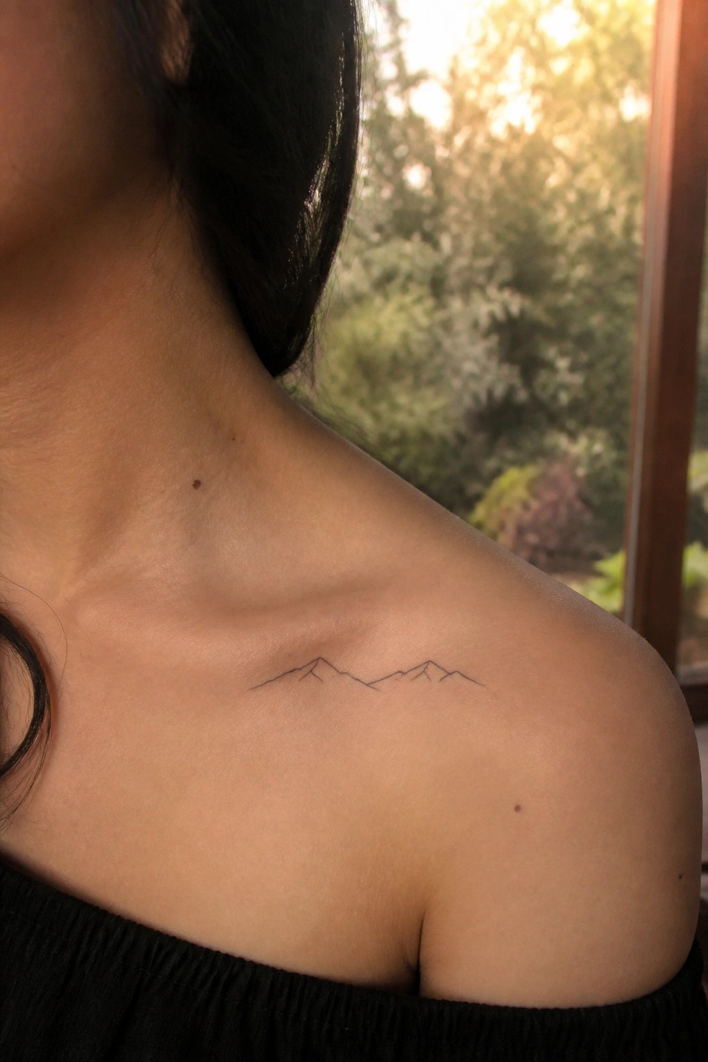 Woman in black off-shoulder top shows minimalist mountain tattoo on shoulder