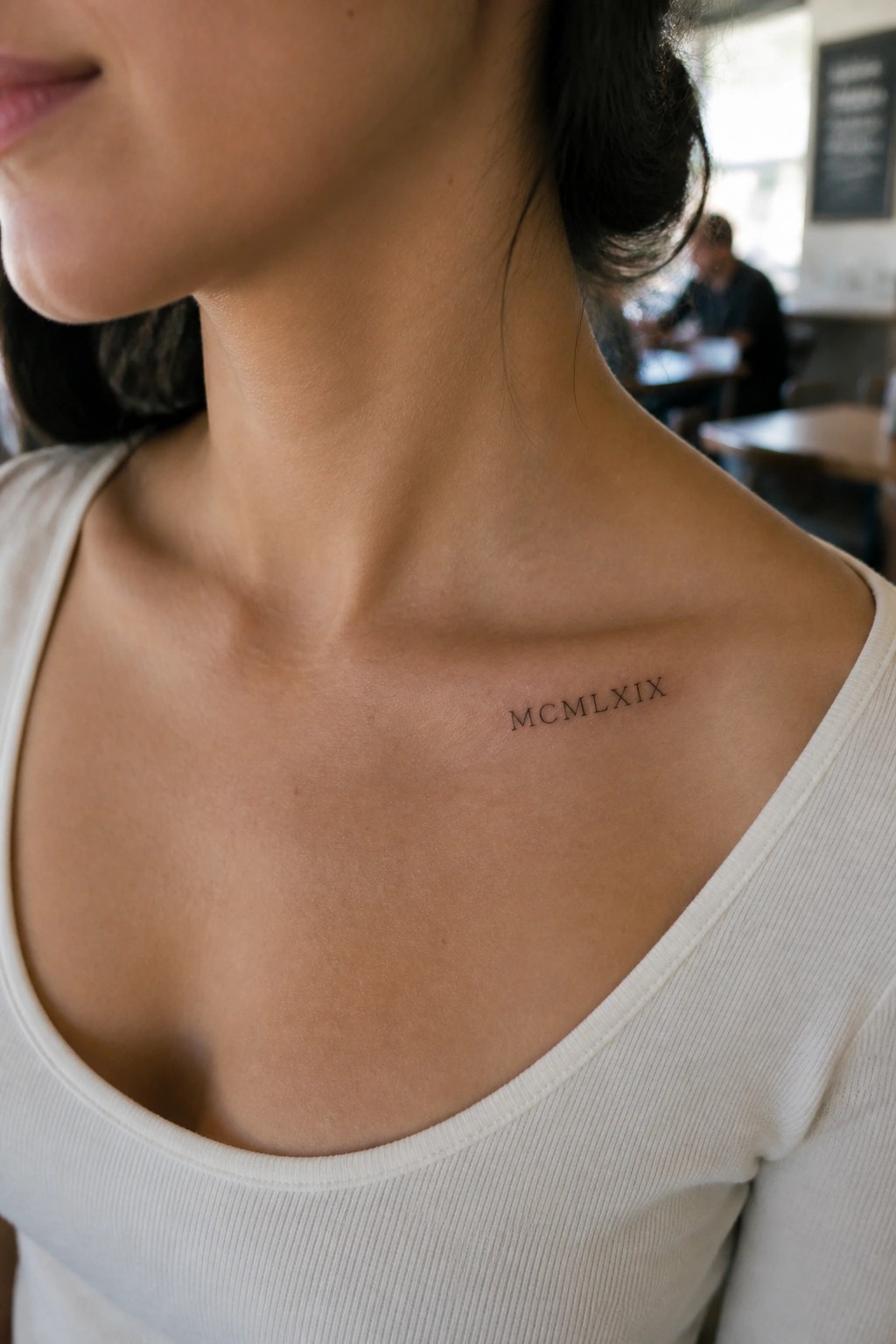 Woman in white top with MCMLXIX tattoo on collarbone in cafe